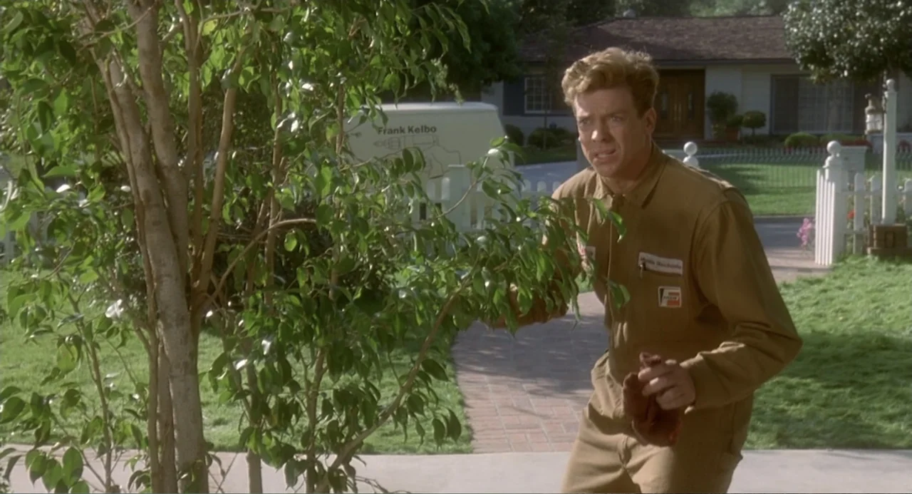 Fatal Instinct (1993) movie still: medium — A suburban front yard features a manicured lawn, a young tree, and a house. A man in a tan work uniform…