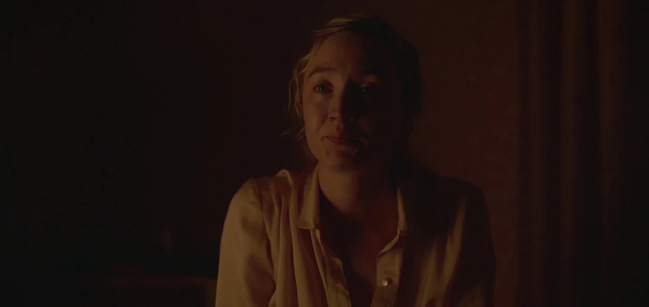 Foe (2023) movie still: medium — A sparsely lit interior space with a dark, indistinct background. A young woman with blonde hair sits facing the…