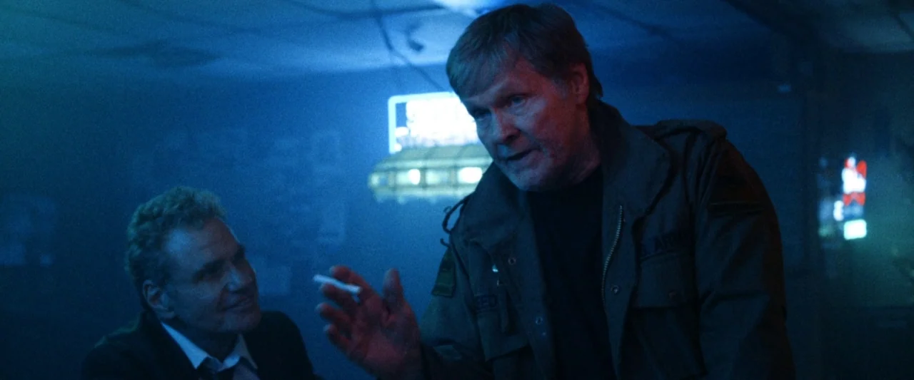 VFW movie still: medium — A dimly lit room with dark walls and a bright glowing neon sign visible behind. Two men stand in the foreground engaged in…
