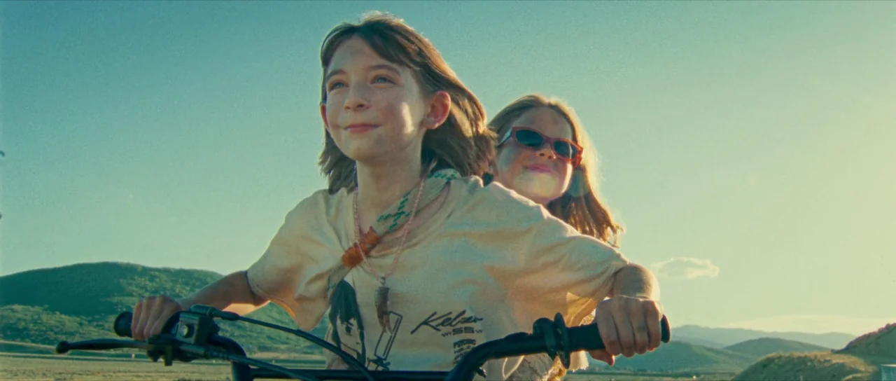 Riddle of Fire (2023) movie still: medium — A rural landscape with rolling green hills under a bright, clear sky. Two young girls ride a bicycle…