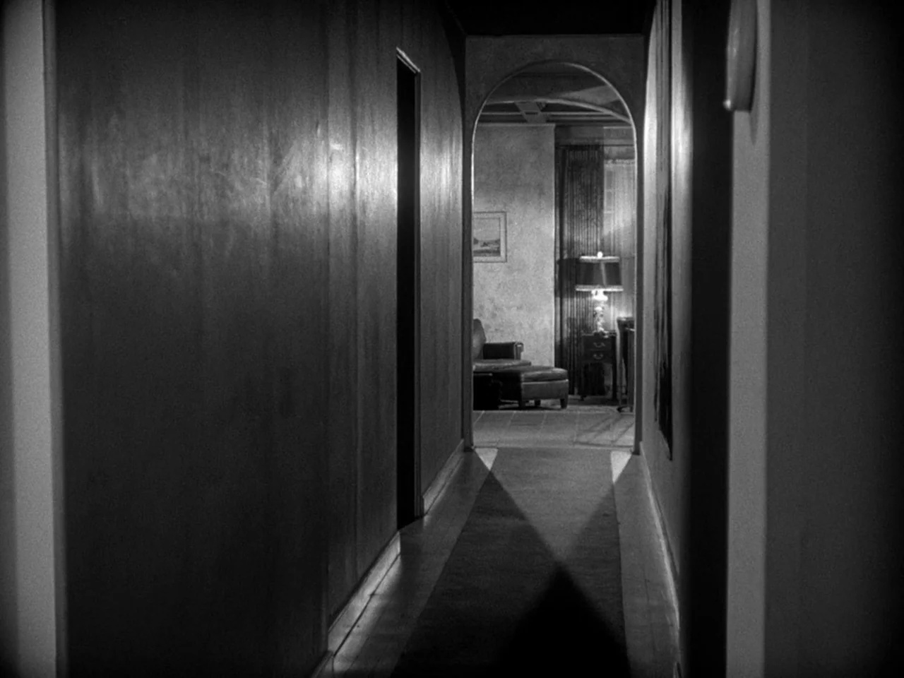 The Prowler (1951) movie still: establishing — A long, dark hallway leads to an illuminated room featuring an arched doorway. A table lamp casts a…