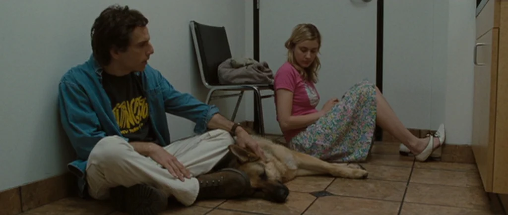 Greenberg (2010) movie still: medium — A clinical waiting area with tan floor tiles and plain white walls. A man sits on the floor petting a dog…