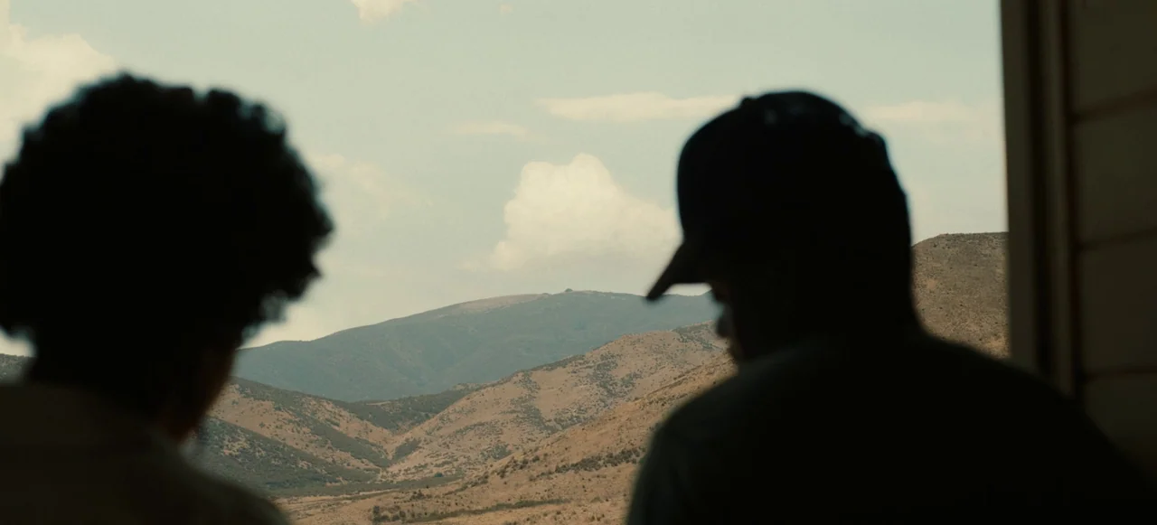 Nope (2022) movie still: medium — A dry, mountainous region with sparsely vegetated hills extends into the distance under a bright sky. Two figures…