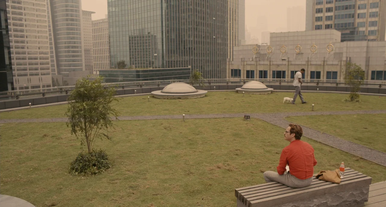 Her (2013) movie still: wide — A green rooftop terrace is surrounded by dense modern skyscrapers in a hazy atmosphere. One man sits on a bench in…