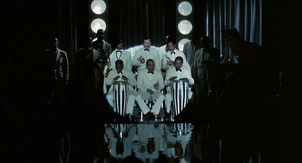 Le Cercle Rouge (1970) movie still: wide — A jazz band stands on a dark stage with circular lighted columns behind them. The musicians play drums…