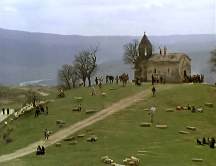The Wishing Tree (1977) movie still: establishing — A stone chapel sits atop a rolling green hillside under a cloudy sky. Many people congregate…