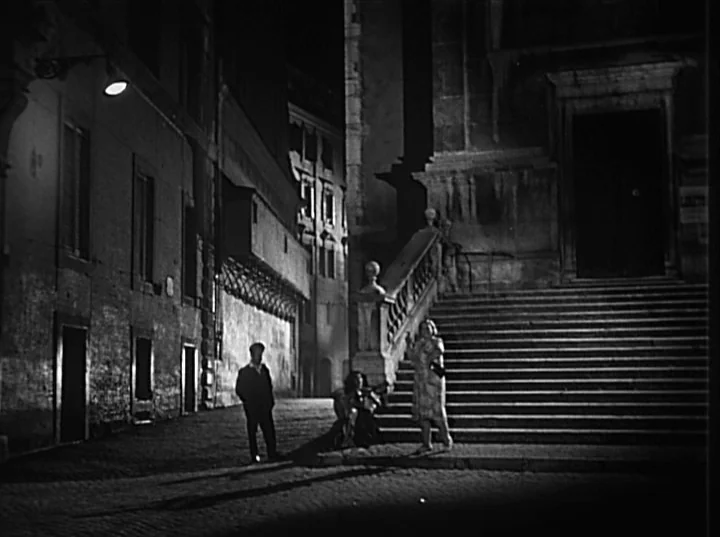 Variety Lights (1950) movie still: wide — A dark, empty city street at night features stone architecture and an ornate staircase. Three individuals…