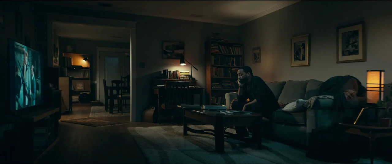 The Life of Chuck (2024) movie still: wide — A dimly lit home interior with a dark living room and an adjacent dining area. A man sits alone on a…
