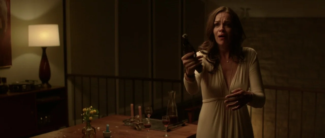 The Invitation (2015) movie still: medium — A dimly lit room features a wooden dining table with glassware and a lamp. A woman in a light dress…