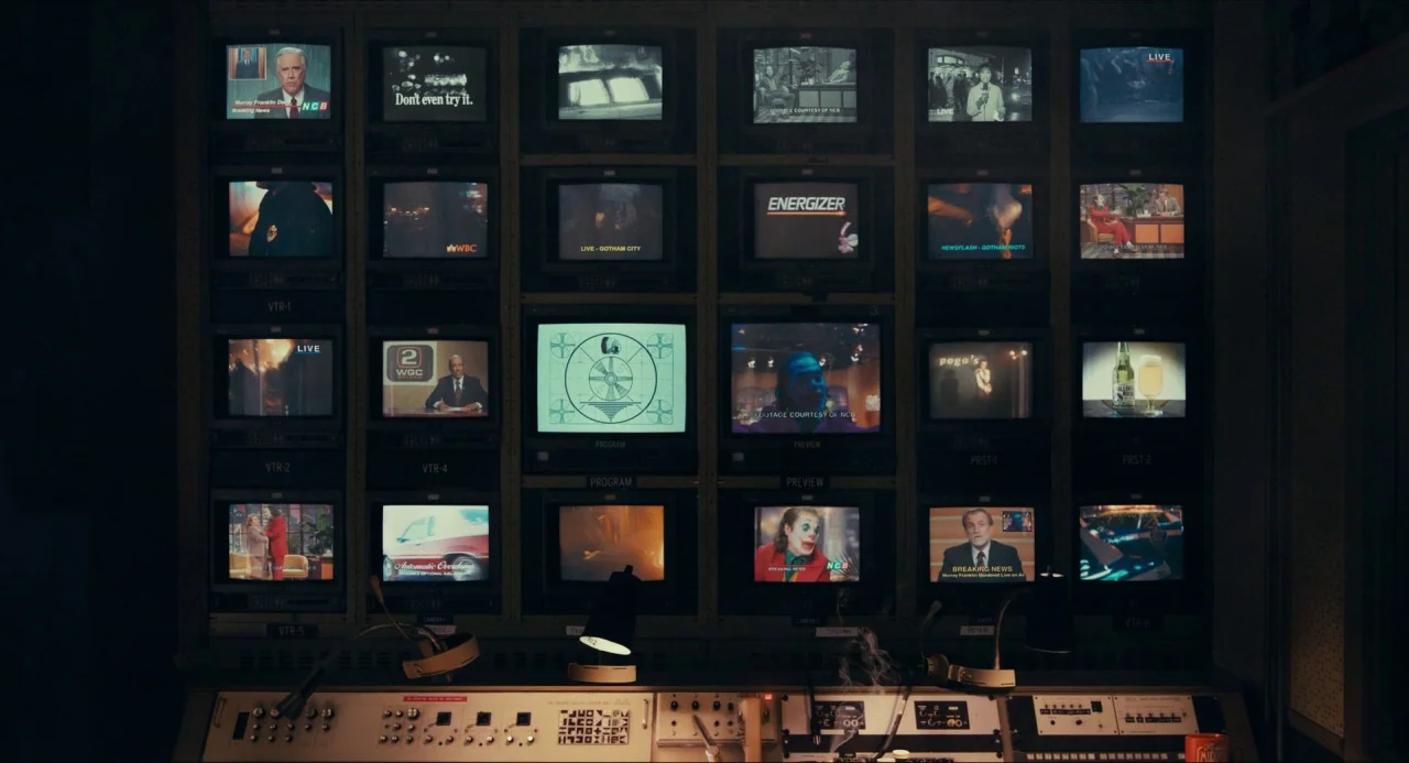 Joker (2019) movie still: wide — A dimly lit broadcast control room features a wall grid of small television monitors. The monitors display news…