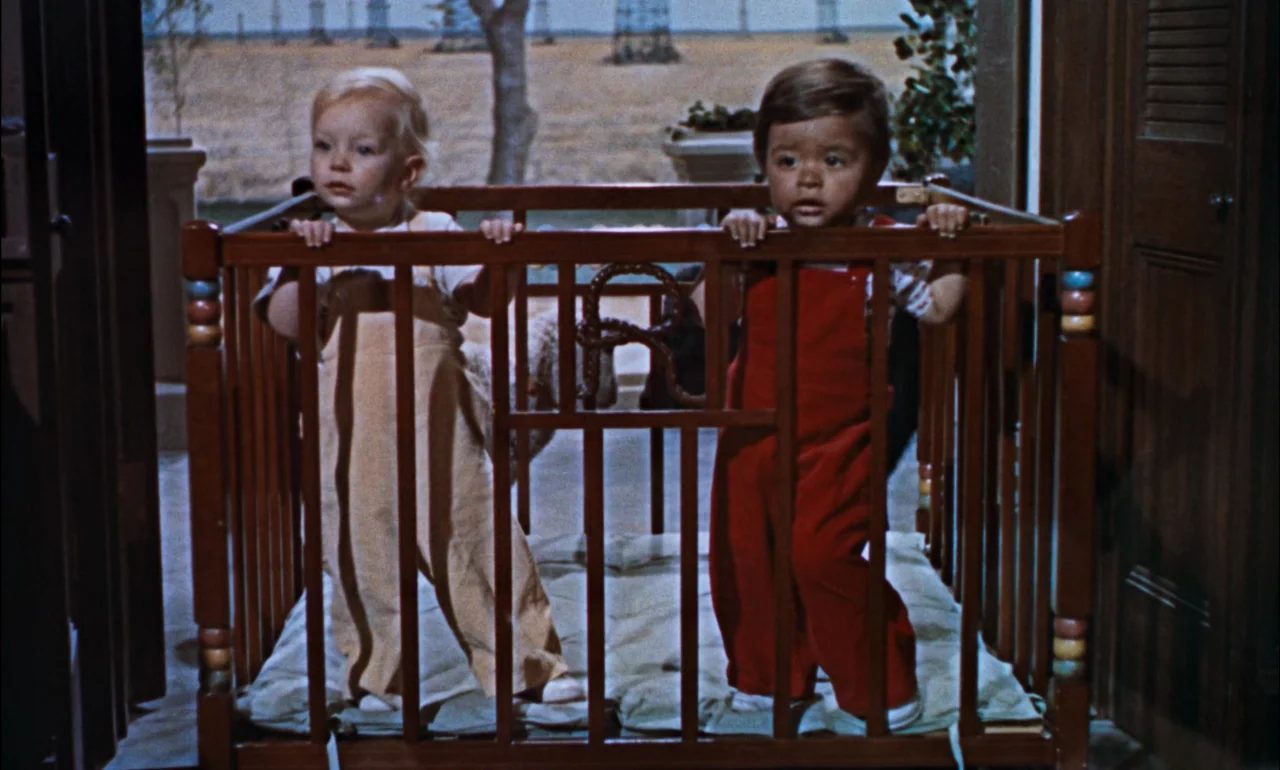 Giant (1956) movie still: medium — Two toddlers stand in a wooden playpen on a covered porch. In the background, a flat field with electrical pylons…