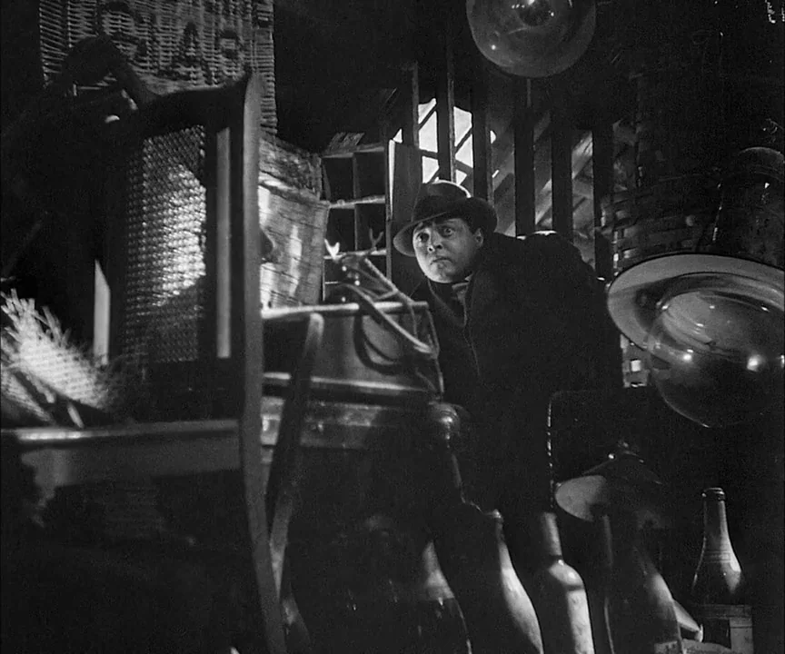 M (1931) movie still: medium — A dark, cluttered space filled with wicker furniture and scattered debris. A man wearing a fedora stares intently…