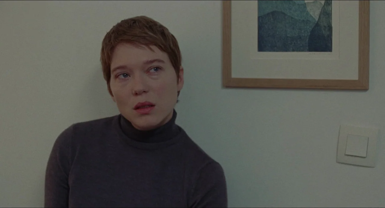One Fine Morning (2022) movie still: medium — A plain interior wall features a framed landscape print and a light switch. A woman stands in front of…