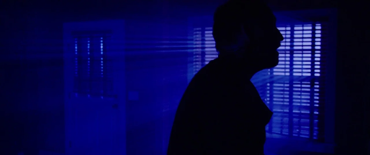 The Mind’s Eye (2015) movie still: medium — A dark interior room is illuminated by thin, horizontal beams of bright blue light. A man stands in…