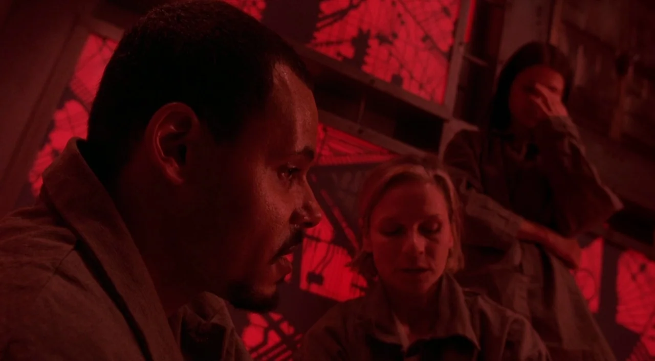 Cube (1997) movie still: medium — A dark enclosure features metallic walls lit with vibrant red geometric patterns. Three people dressed in…