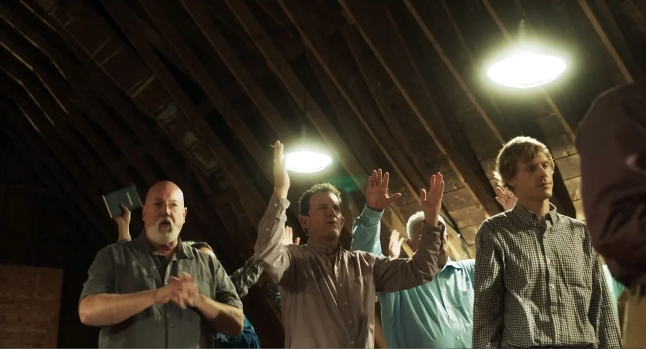 Salvation (2025) movie still: medium — An attic with visible wooden ceiling rafters and two bright overhead pendant lights. Several men stand in a…