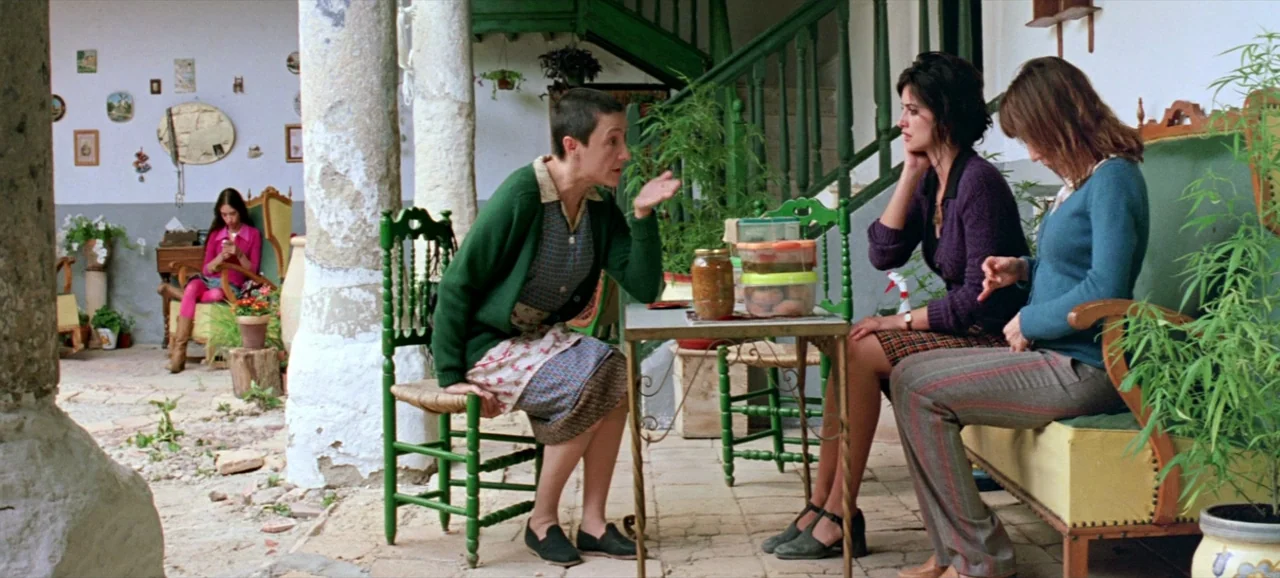 Volver (2006) movie still: wide — A rustic Spanish courtyard features whitewashed stone columns and green painted wooden stairs. Three women are…