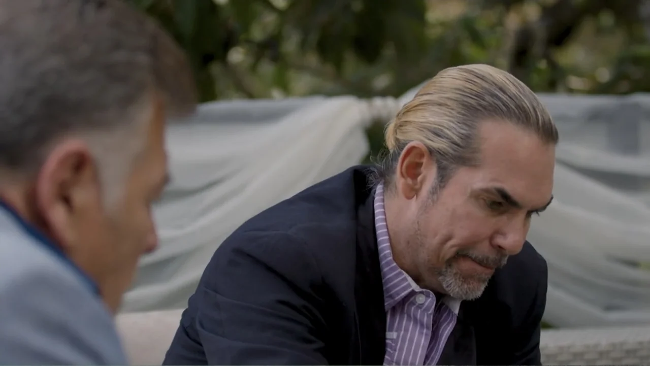 Por Amor al Money (2023) movie still: medium — An outdoor patio setting features white fabric draping and blurred greenery in the background. A man…