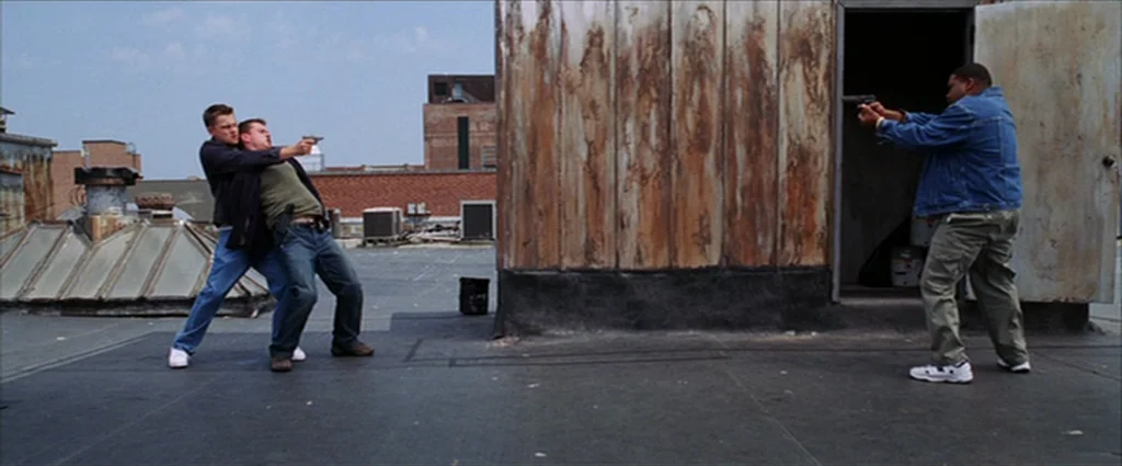 The Departed (2006) movie still: wide — An urban roof with corrugated metal walls and distant brick buildings under clear sky. Two men interact in a…