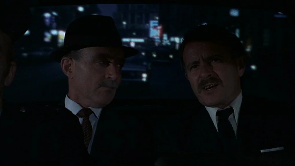 Frenzy (1972) movie still: medium — The interior of a moving car at night with a blurred city background. Two men in formal business suits sit side…