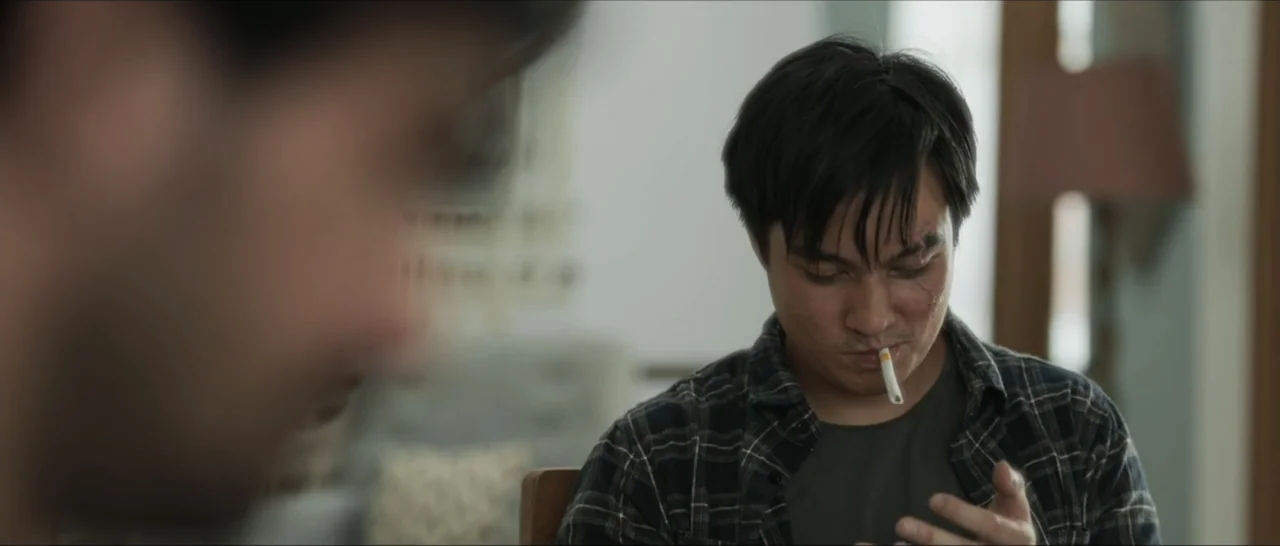Innocent Vengeance (2023) movie still: medium — An interior space features a man sitting in the foreground of a blurred room. The man holds a lit…