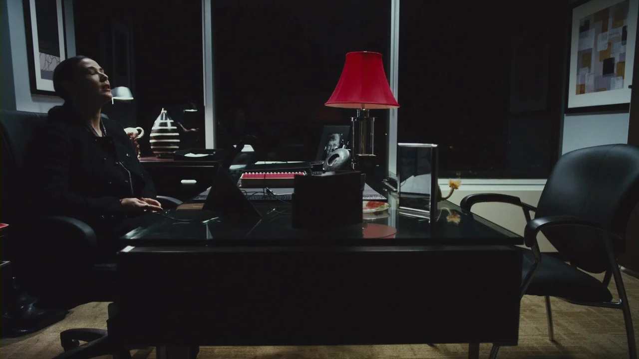 Margin Call (2011) movie still: medium — A dark office at night features a glass desk and a red-shaded table lamp. A woman sits in a chair with her…