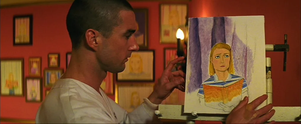 The Royal Tenenbaums (2001) movie still: medium — A man stands before an easel in a room with numerous framed drawings on red walls. He gestures…