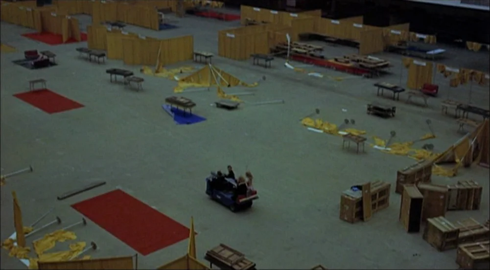 The King Of Marvin Gardens movie still: wide — A vast, industrial hall contains numerous scattered wooden partitions, tables, and red floor mats. A…