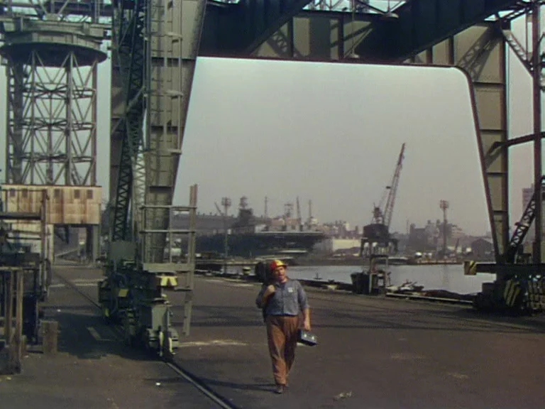 On The Town (1949) movie still: wide — A massive gantry crane dominates a wide industrial shipyard near the water. A man in work clothes walks…
