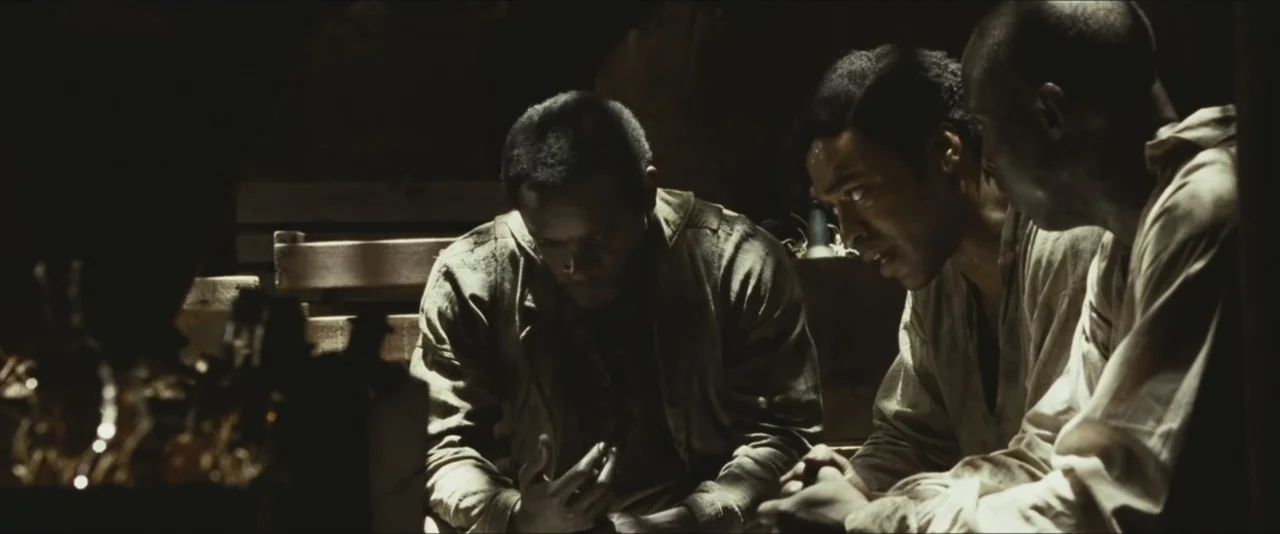 12 Years a Slave (2013) movie still: medium — A dark, cramped, wooden-walled enclosure serves as the setting for three men talking. The men are…