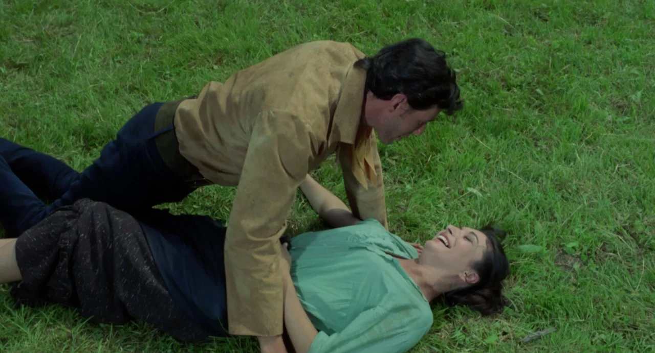 Johnny Colt (1966) movie still: medium — A man and woman lie on a patch of bright green grass. The man looms over the woman while she laughs toward…