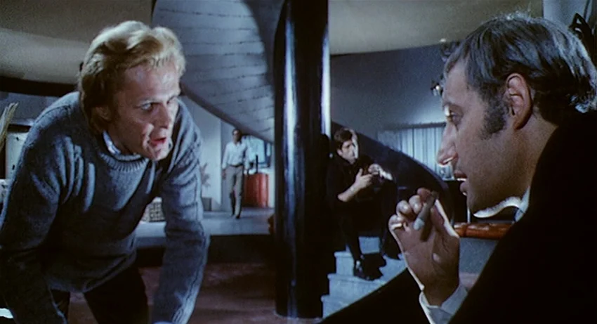 5 Dolls for an August Moon (1970) movie still: medium — A modernist interior features a curved central column and a sunken lounge area. One man…