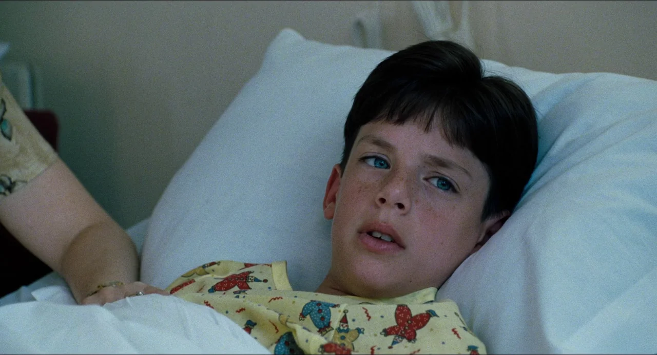 Happiness (1998) movie still: closeup — A white sterile environment featuring a hospital bed with a white pillow and sheet. A young boy with…