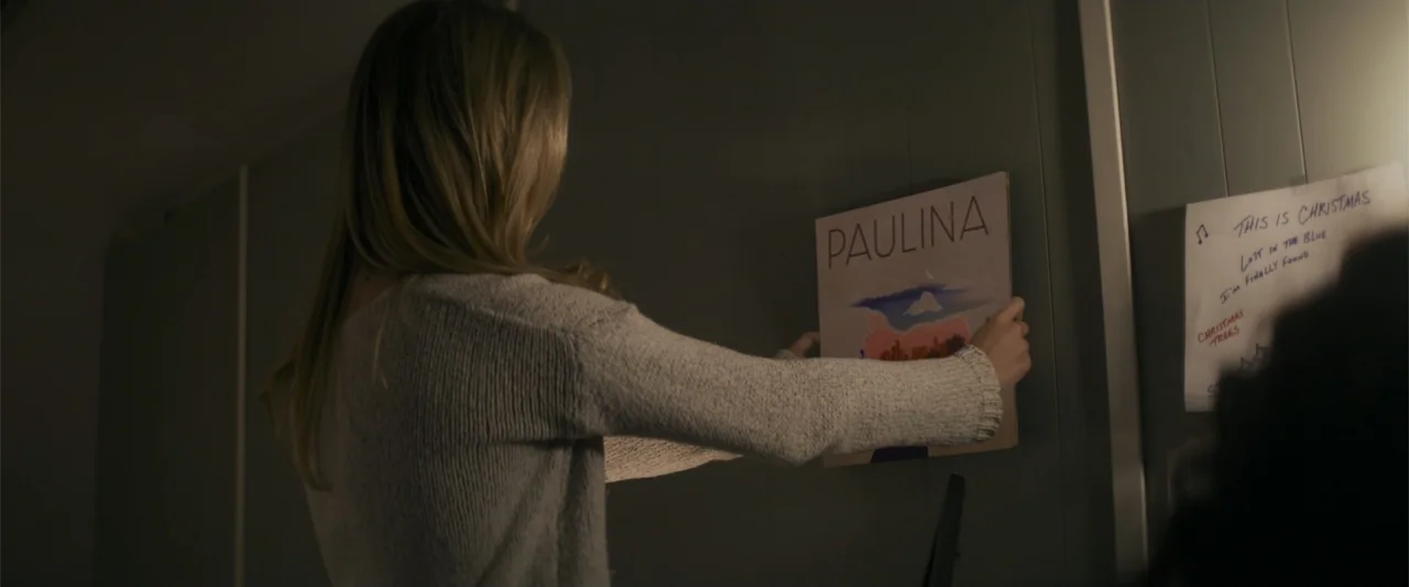 Holiday Harmony (2022) movie still: medium — An interior space features pale grey paneled walls and a hand-written note on paper. A young woman in a…