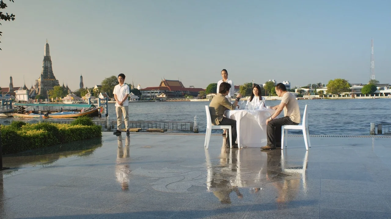 Zero Tolerance (2015) movie still: wide — A stone-paved riverside terrace overlooks a wide river with temples visible on the far bank. Four people…