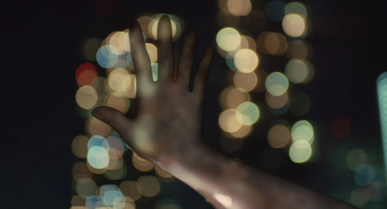 Air Doll (2009) movie still: closeup — A dark night setting filled with blurry, glowing circular bokeh lights from a city. A human hand is…