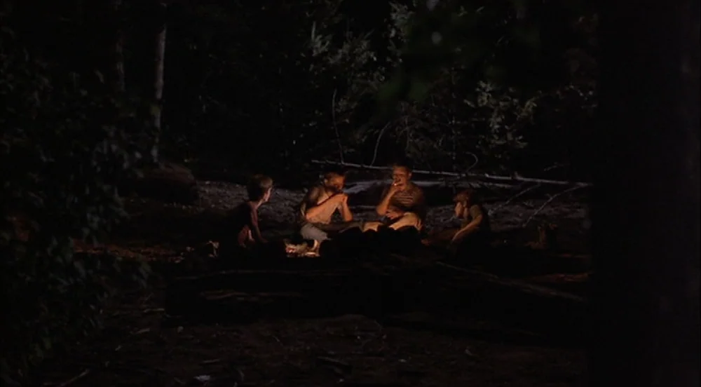 Stand By Me (1986) movie still: wide — A group of four teenage boys sits on the dark ground around a campfire. The surrounding area consists of…
