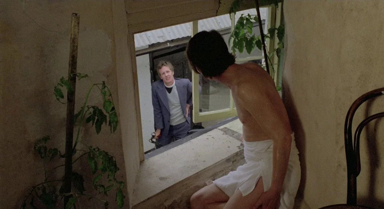 The Shout (1978) movie still: medium — A man in a towel sits inside near an open window while another man stands outside. A bicycle handle and…
