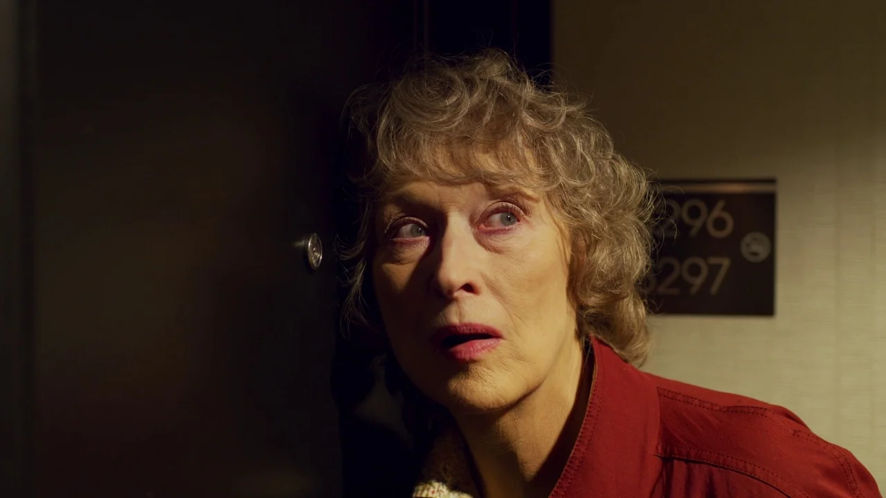 The Laundromat (2019) movie still: closeup — A dimly lit hotel corridor features a room door with a metal peephole. An older woman listens closely…