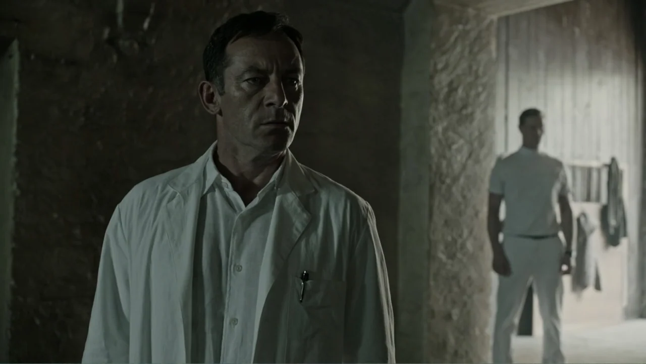 A Cure for Wellness (2017) movie still: medium — A stone-walled medical room with a dark corner and a brightly lit background area. A doctor in a…