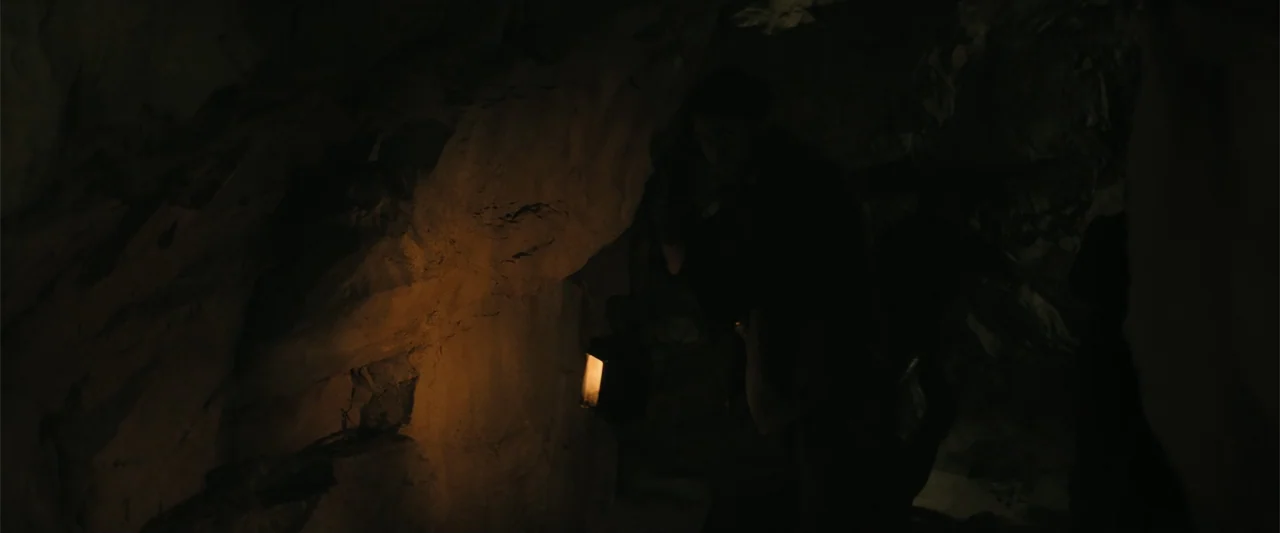 The Bluff (2026) movie still: medium — A dark subterranean cave with rough stone walls and narrow passages. A person stands near a wall holding a…