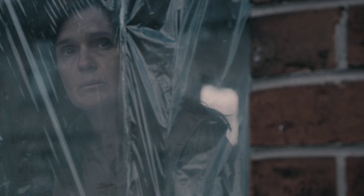 The Selfish Giant (2013) movie still: closeup — A woman stands behind a clear, wrinkled plastic sheet next to a weathered brick wall. She looks…