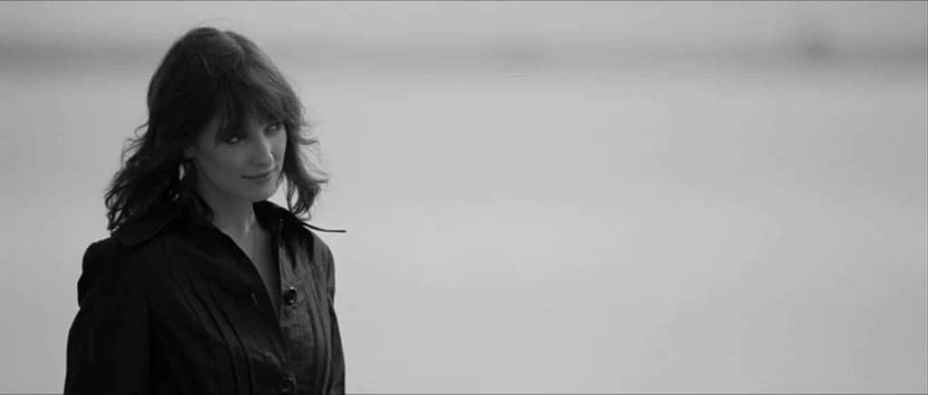 Control movie still: medium — An open, light-filled environment with a flat, bright background suggestive of a beach. A woman with dark hair and a…