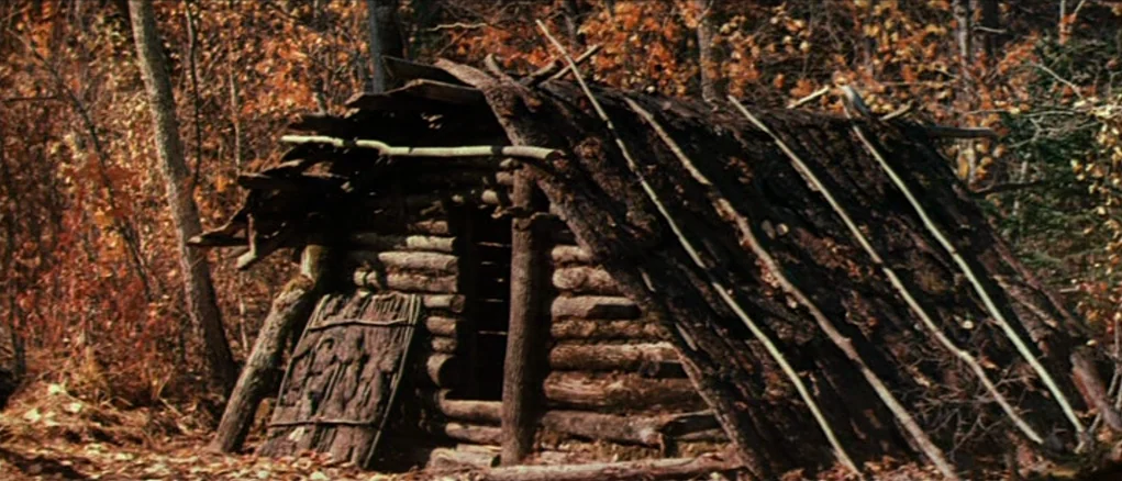 Dersu Uzala (1975) movie still: wide — A rustic log structure with a rough timber roof sits in an autumn forest. Fallen leaves blanket the ground…