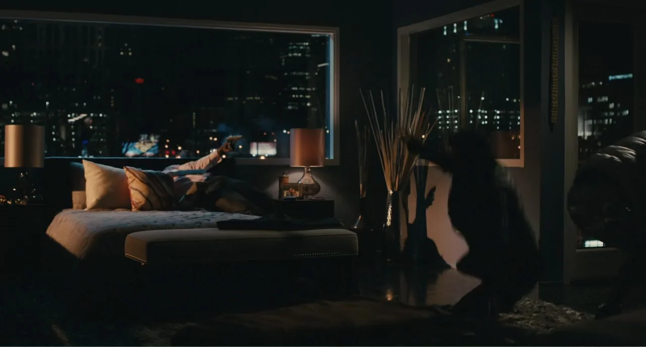 Lila & Eve (2015) movie still: wide — A dark penthouse bedroom features large windows overlooking a city skyline at night. One man aims a firearm…