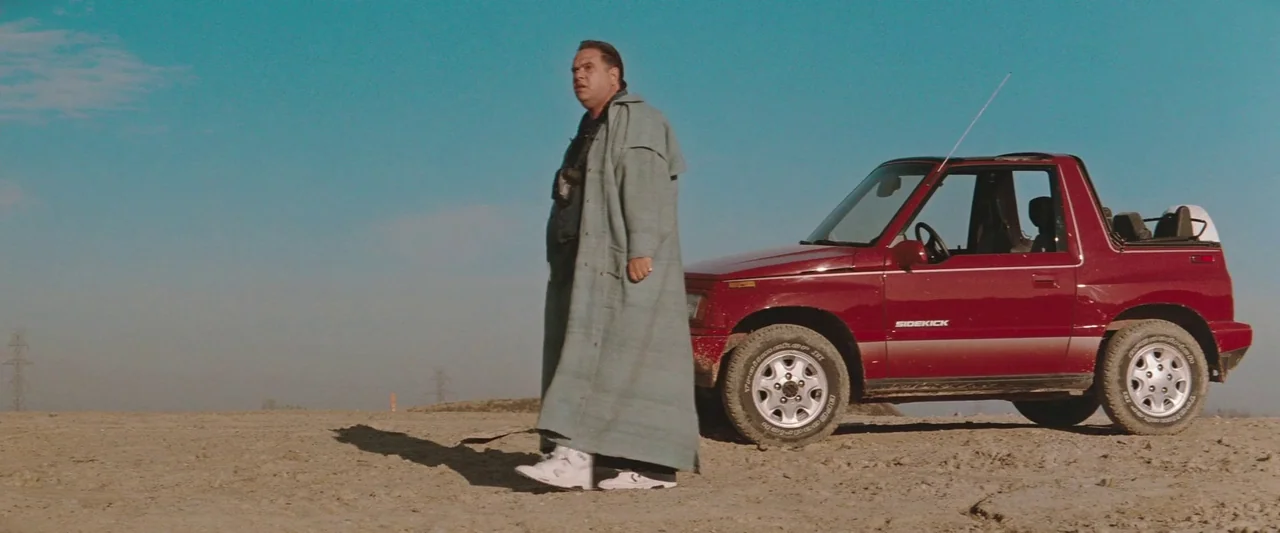 The Adjuster (1991) movie still: fullbody — A man in a long light-colored coat walks across a dry, dusty dirt lot. A red compact SUV is parked…
