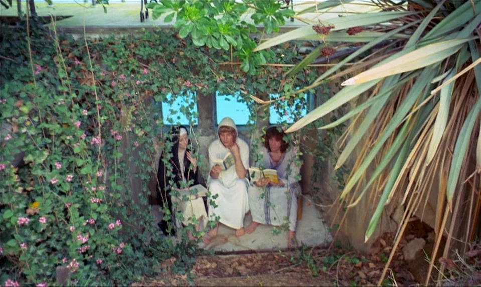 Lions Love (1969) movie still: wide — A stone alcove overgrown with ivy and flowering vines is set into the earth. Three people sit on the ground…