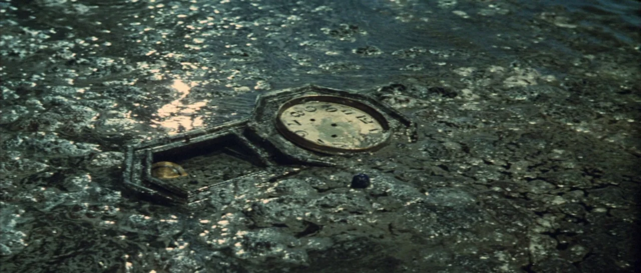 Godzilla Vs Hedorah (1971) movie still: closeup — A dark muddy bank of a marsh features a partially submerged vintage clock face. The antique…