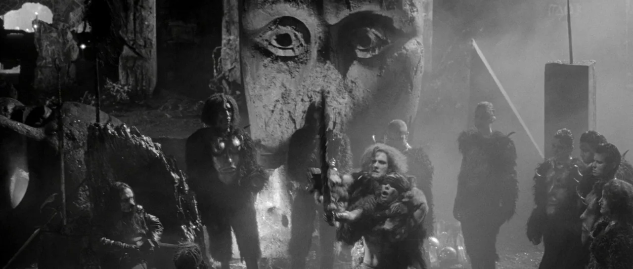 She Is Conann (2023) movie still: wide — A dark stone temple features a massive sculpted face embedded in the rear wall. A group of individuals…