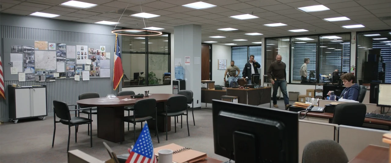 Trap House (2025) movie still: wide — A busy open-plan office space contains desks, cubicles, and a conference table area. Several men in work…
